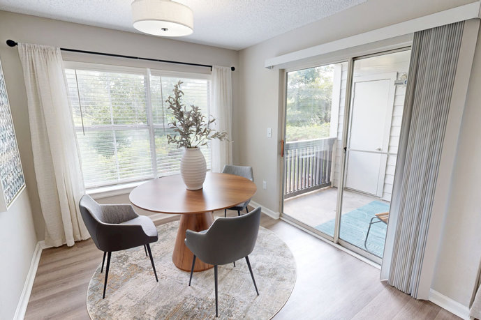 Beige apartment dining area that seats 4 with a sliding glass door to the right and door to the bedroom on the left